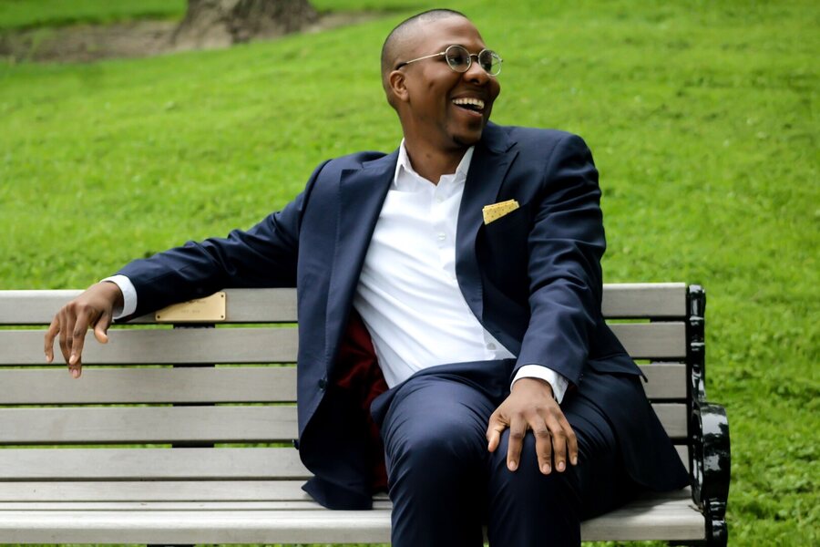 Gabriel Walker leaning back on a park bench in a navy suit, smiling broadly.